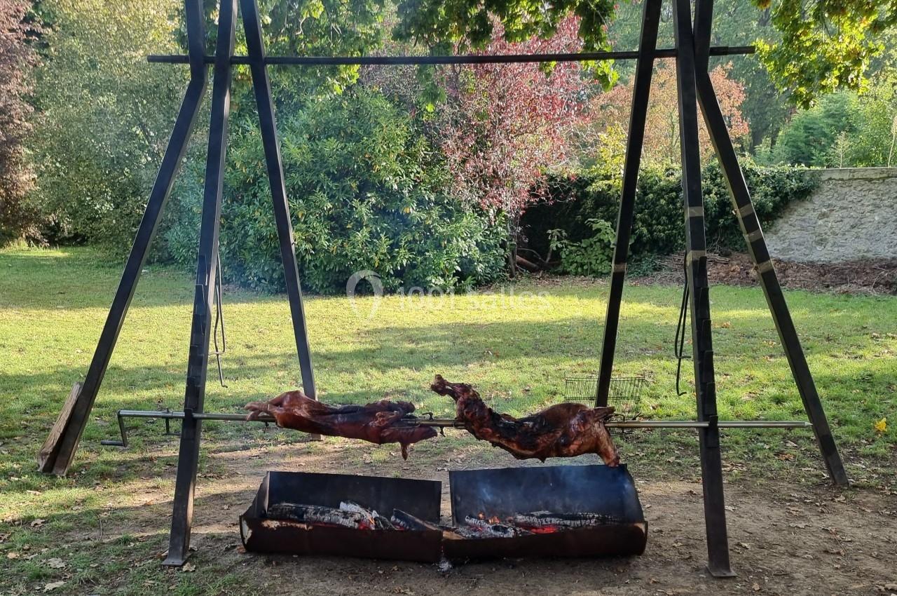 Deux porcelets rôtissent sur une broche au-dessus de braises dans un cadre extérieur verdoyant.