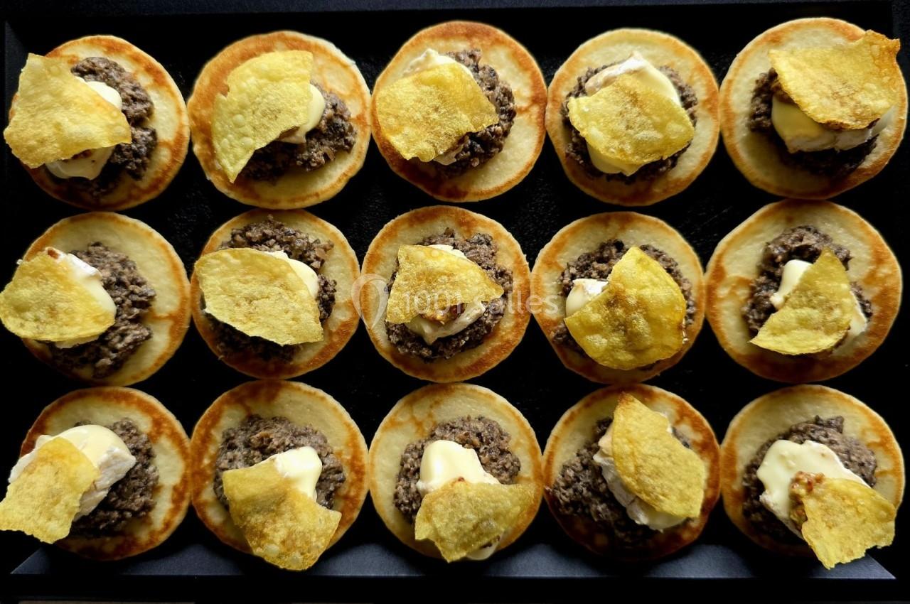 Mini-blinis garnis de viande hachée, crème et chips dorées, disposés en rangées sur un plateau noir.