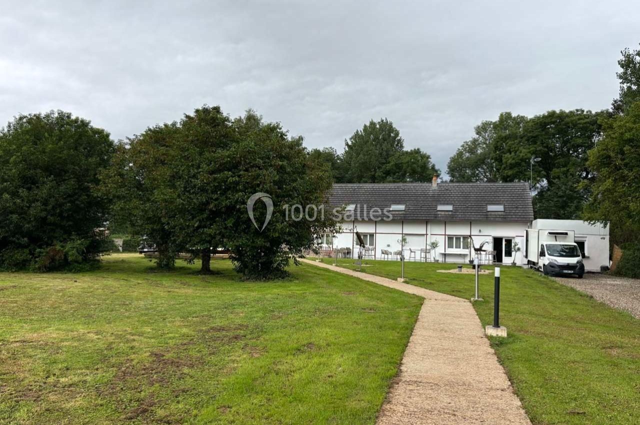 Chemin en béton menant à une maison blanche entourée d'arbres et d'une pelouse sous un ciel nuageux.