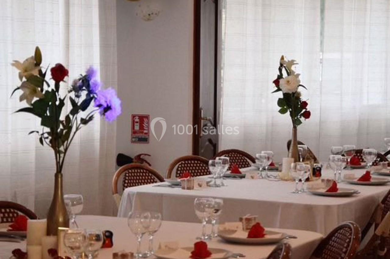 Salle de réception avec tables dressées, nappes blanches, verres, serviettes rouges et vases de fleurs.
