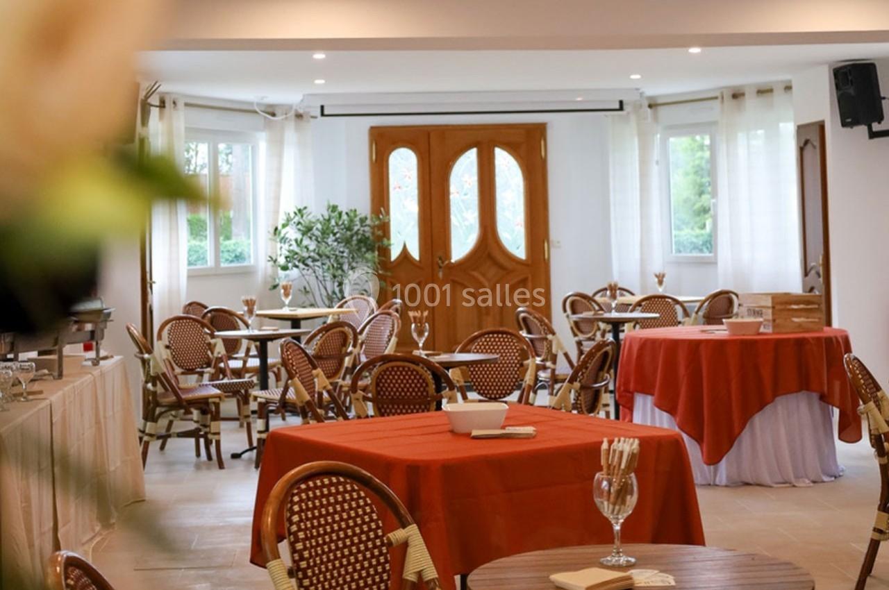 Salle lumineuse avec tables rondes et chaises en osier, nappes rouges et blanches, et grandes fenêtres donnant sur l…
