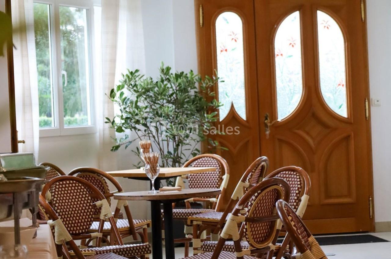 Salle lumineuse avec chaises en rotin, tables rondes, plante verte et porte en bois avec vitraux décoratifs.