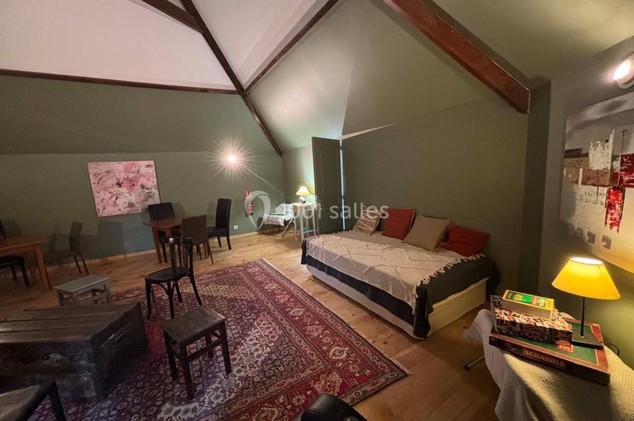 Grande pièce mansardée avec lit, tables, chaises, tapis coloré et éclairage tamisé, décorée de tableaux modernes.