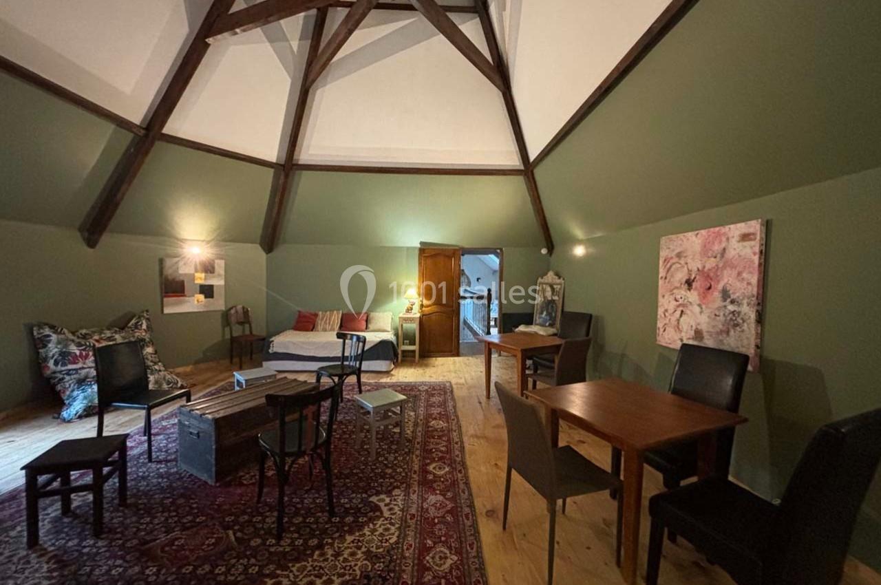 Salle mansardée avec poutres apparentes, mobilier en bois, tapis coloré, et éclairage tamisé.