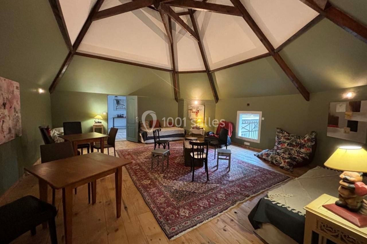 Salle mansardée avec poutres apparentes, tapis au sol, mobilier varié et éclairage chaleureux.