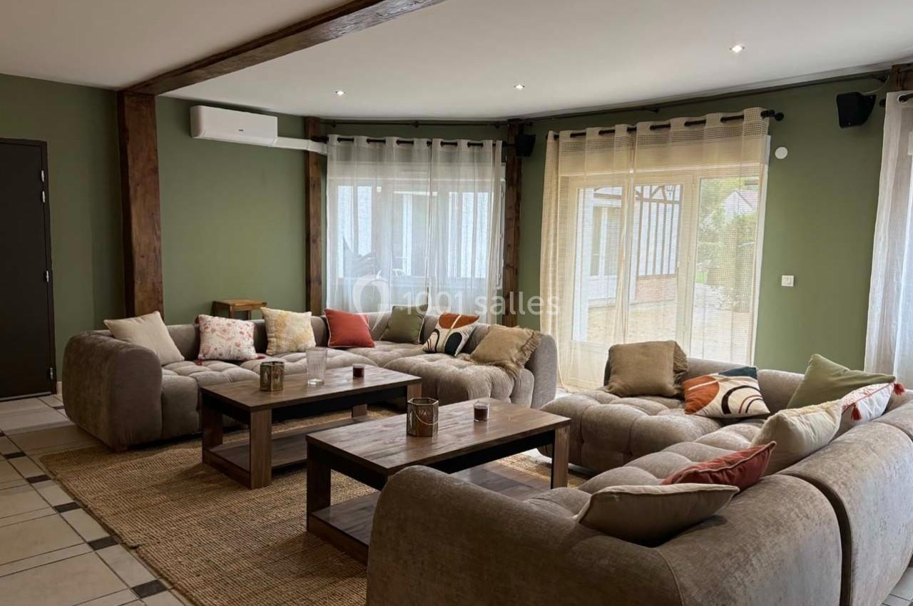 Salon spacieux avec canapés beiges en angle, tables basses en bois, tapis en jute et grandes fenêtres avec rideaux clairs.