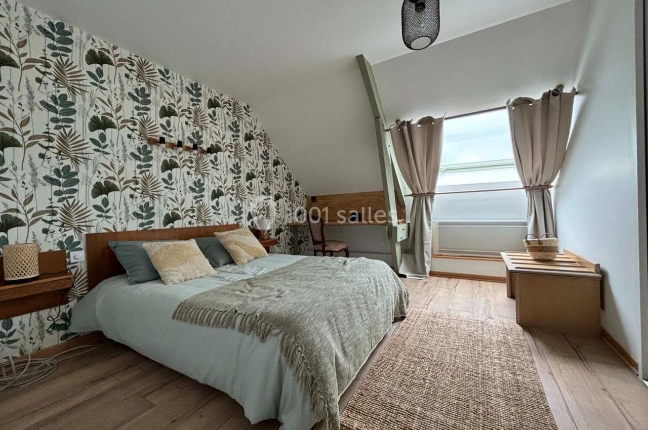 Chambre lumineuse avec lit double, décoration végétale, bureau en bois, tapis en jute et grande fenêtre avec rideaux beiges.