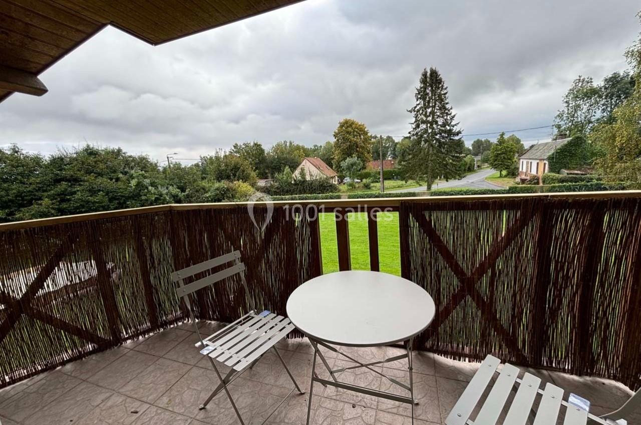 Vue depuis un balcon avec une table ronde, deux chaises blanches et un paysage rural sous un ciel nuageux.