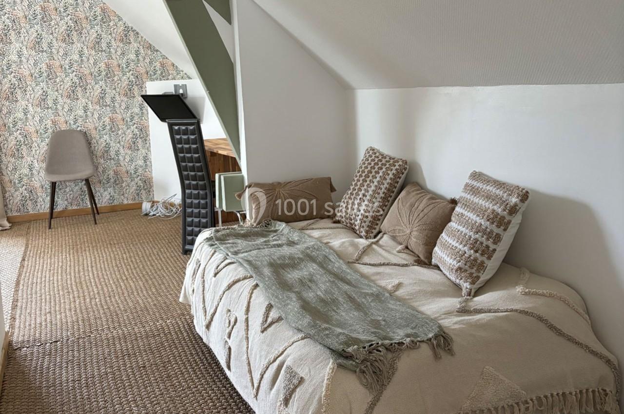 Lit simple avec coussins décoratifs et plaid dans une chambre mansardée au sol en jonc de mer, mur tapissé floral.