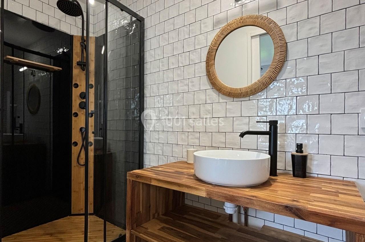 Salle de bain moderne avec douche à paroi vitrée, lavabo rond sur meuble en bois et miroir encadré en rotin.