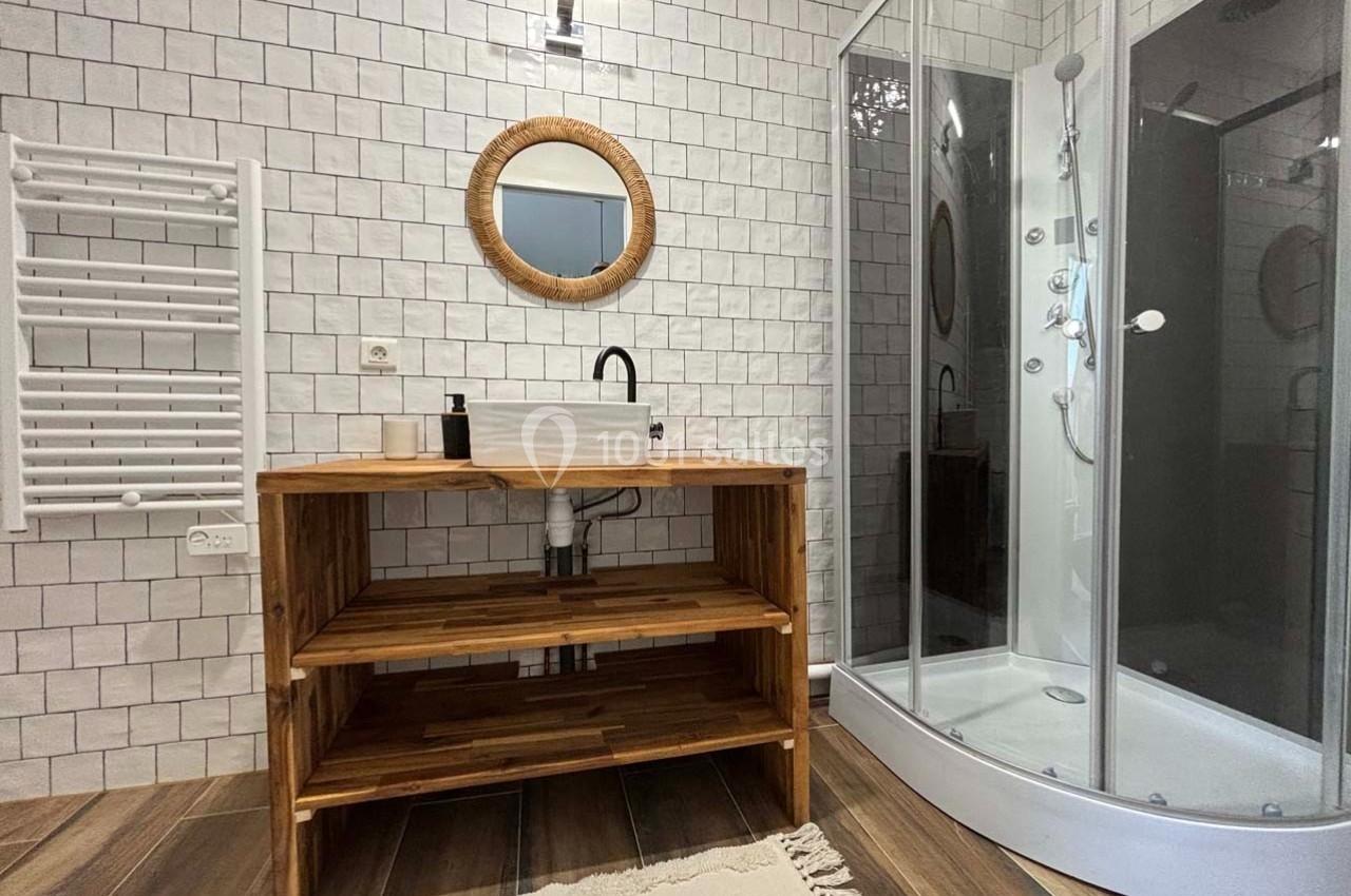Salle de bain avec douche arrondie, meuble en bois avec vasque blanche, miroir rond et carrelage mural blanc.