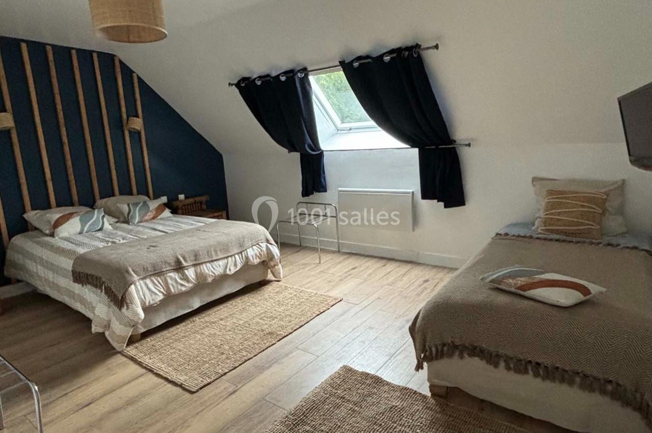 Chambre lumineuse avec un lit double, un lit simple, parquet au sol, tapis en jute et fenêtre avec rideaux noirs.