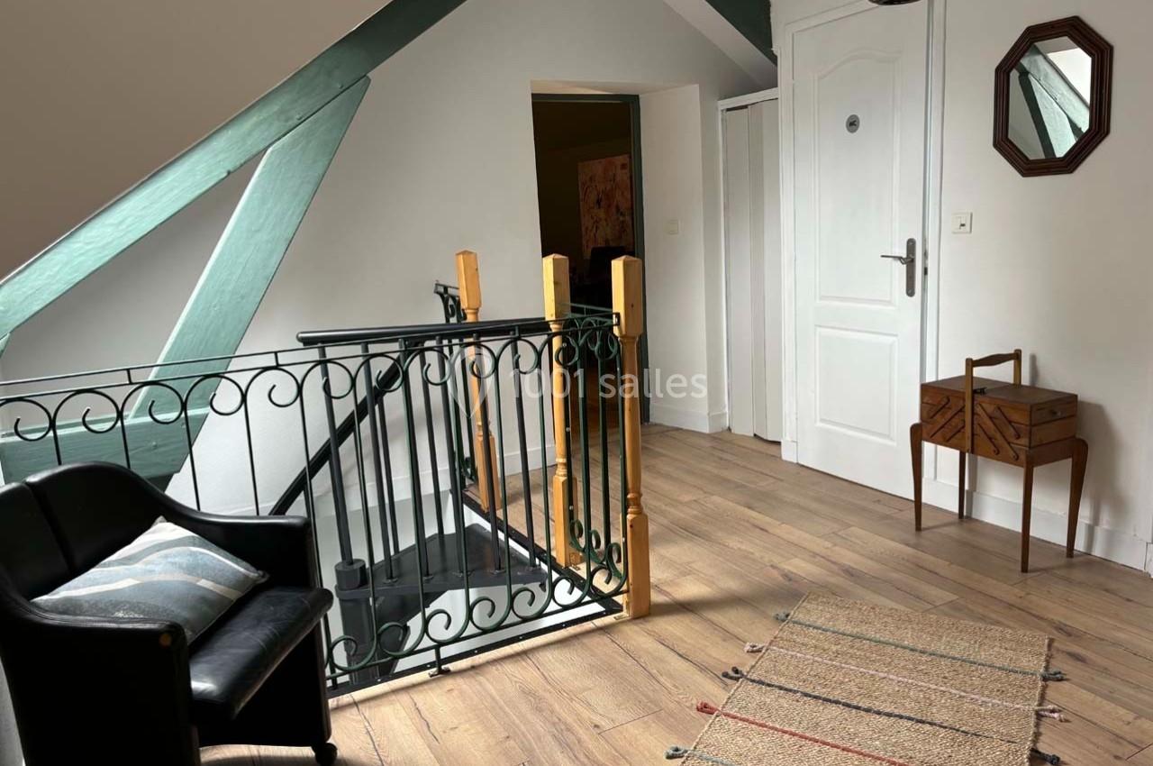 Palier lumineux avec parquet, fauteuil noir, tapis beige, table en bois et escalier entouré d'une rambarde en fer forgé.