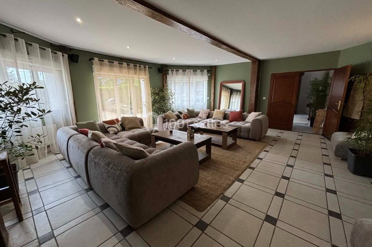 Salon spacieux avec canapés gris, table basse en bois, tapis beige et grandes fenêtres laissant entrer la lumière.