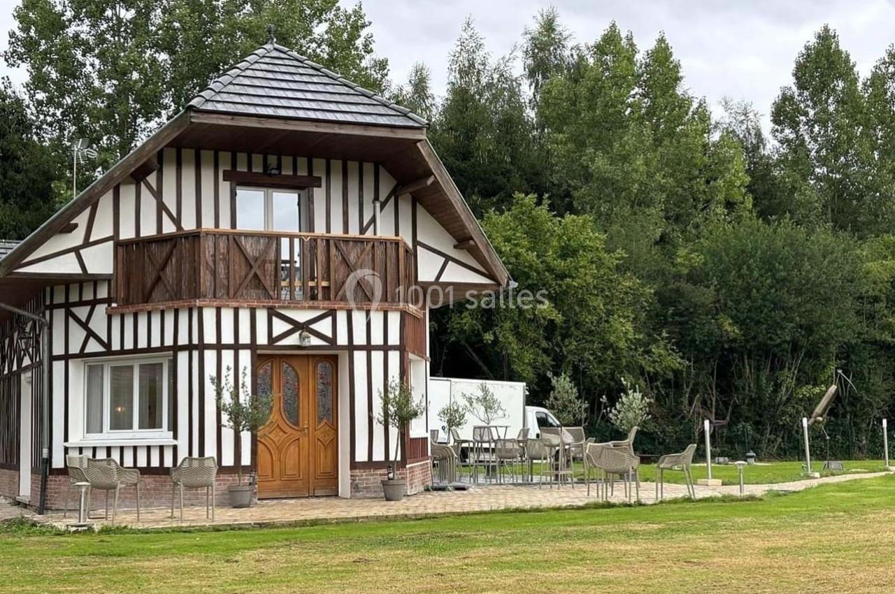 Maison à colombages avec balcon, entourée de chaises et de verdure, située dans un environnement arboré.