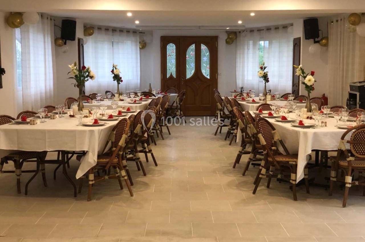 Salle décorée pour un événement, avec des tables dressées, nappes blanches, chaises en rotin et arrangements floraux.