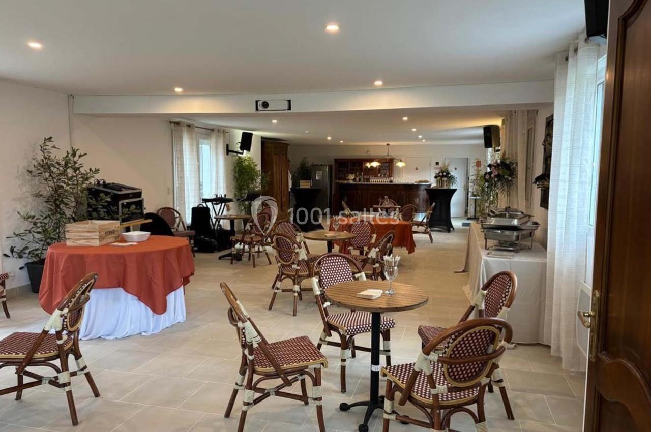 Salle lumineuse aménagée avec des tables et chaises en rotin, un buffet, et un bar en arrière-plan.