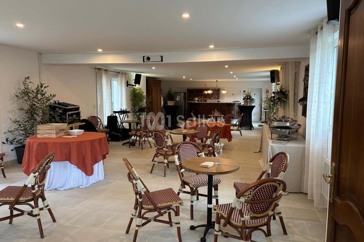 Salle lumineuse aménagée avec des tables et chaises en rotin, un buffet, et un bar en arrière-plan.