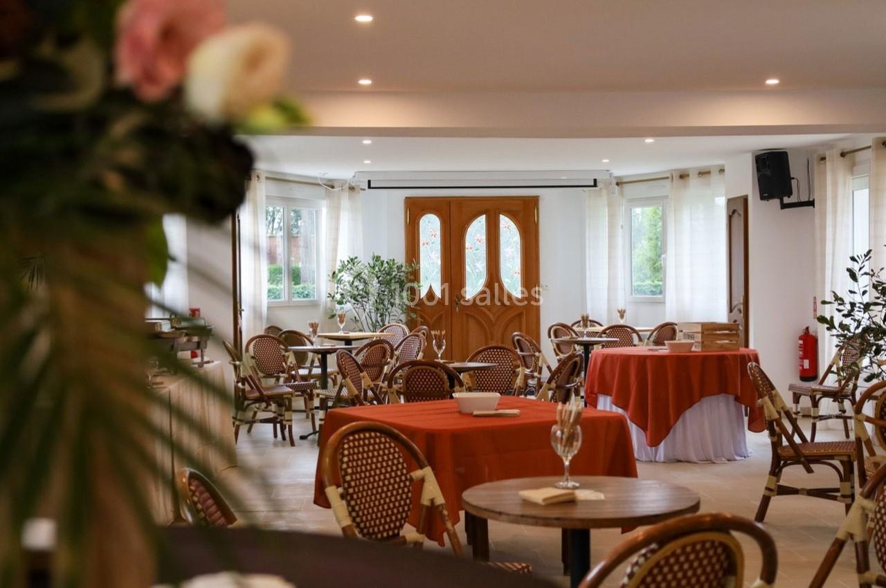Salle lumineuse avec tables rondes et chaises en rotin, nappes orange et blanches, grandes fenêtres et porte en bois sculpté.