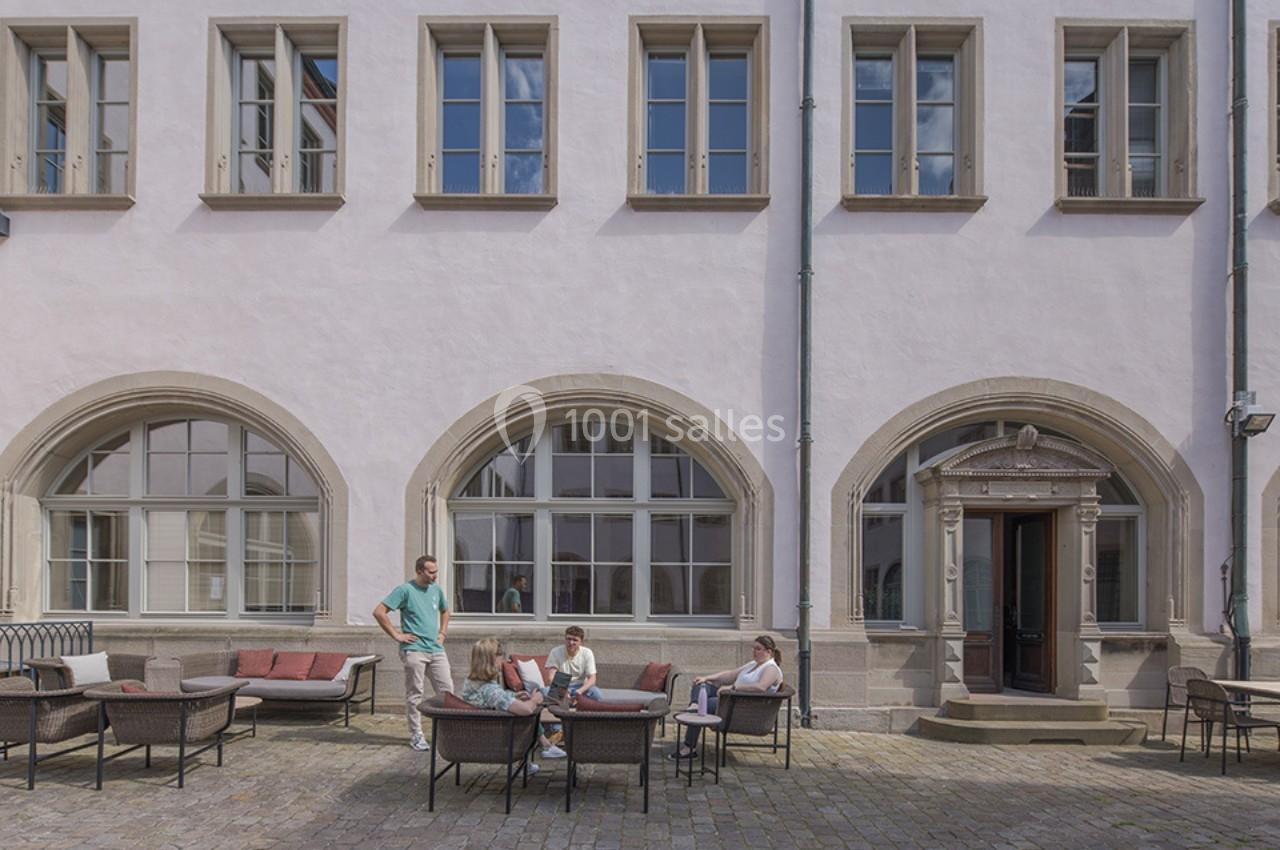 Personnes assises et discutant dans une cour pavée devant un bâtiment aux grandes fenêtres et arches.