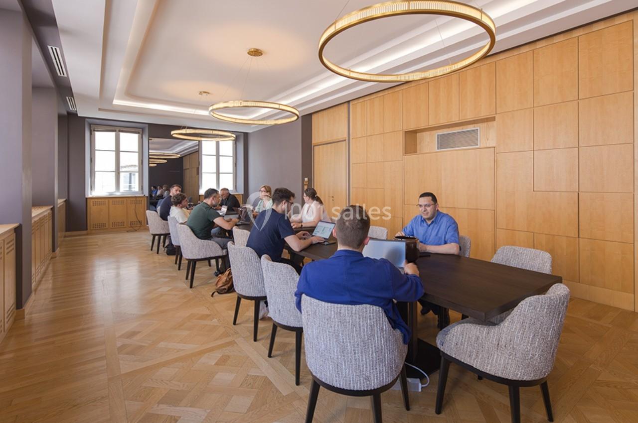Des personnes travaillent autour de deux grandes tables dans une salle lumineuse avec des murs en bois et des lustres…