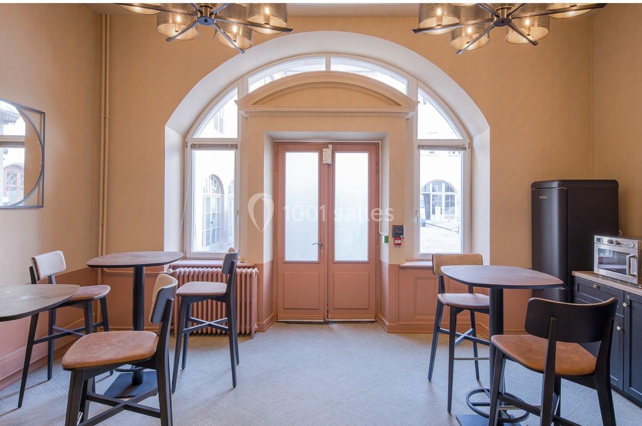 Salle lumineuse avec tables hautes, chaises, grande porte vitrée, fenêtres cintrées et coin kitchenette équipé.