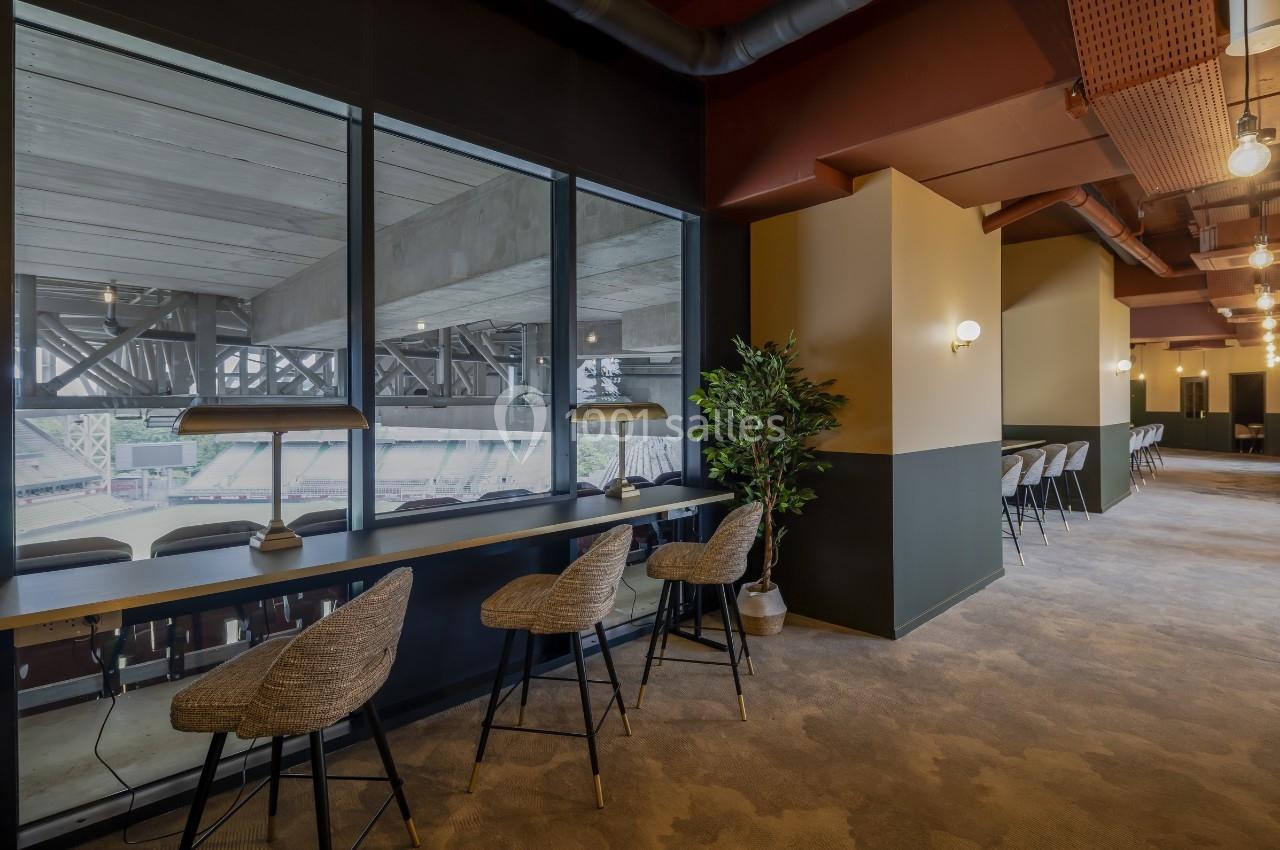 Espace intérieur moderne avec comptoir, chaises en rotin, lampes et vue sur des gradins à travers de grandes fenêtres.
