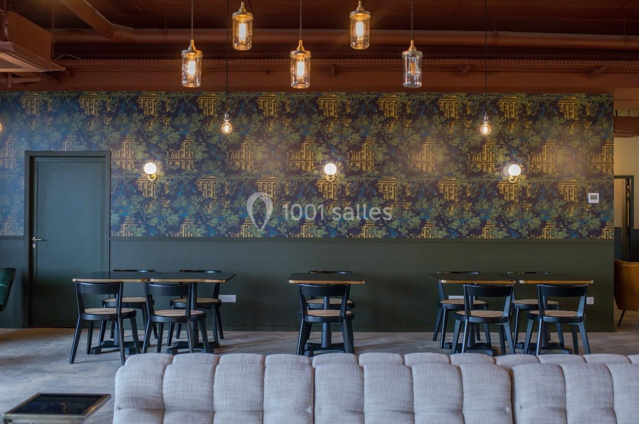 Salle de restaurant avec tables en bois, chaises noires, mur décoré de papier peint floral et luminaires suspendus.