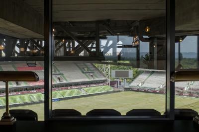 Vue depuis une loge vitrée sur un stade de football vide, avec gradins et pelouse partiellement éclairés.