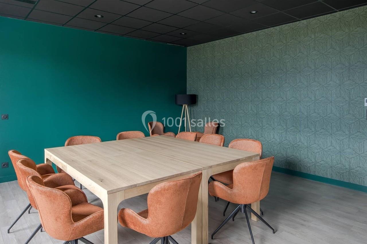 Salle de réunion avec table en bois clair, chaises orange, murs verts et éclairage naturel.