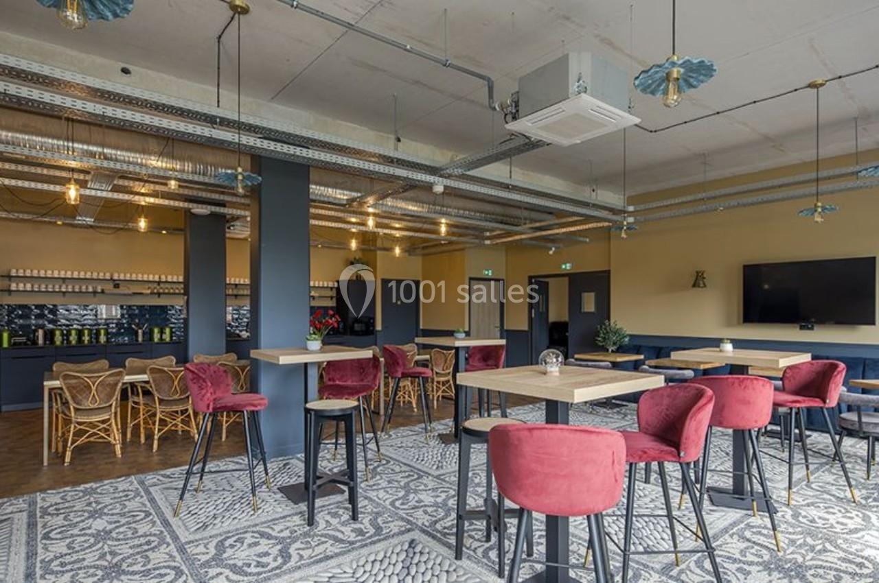 Salle moderne avec tables hautes, chaises rouges, bar en arrière-plan et éclairage suspendu dans un décor industriel.