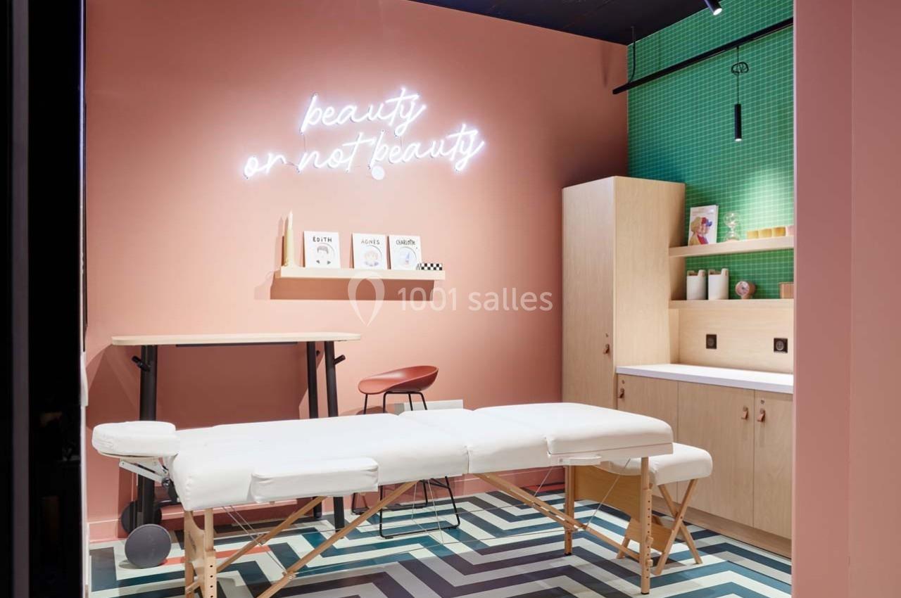 Salle moderne avec table de massage blanche, mur rose avec néon ’beauty or not beauty’ et étagères en bois.