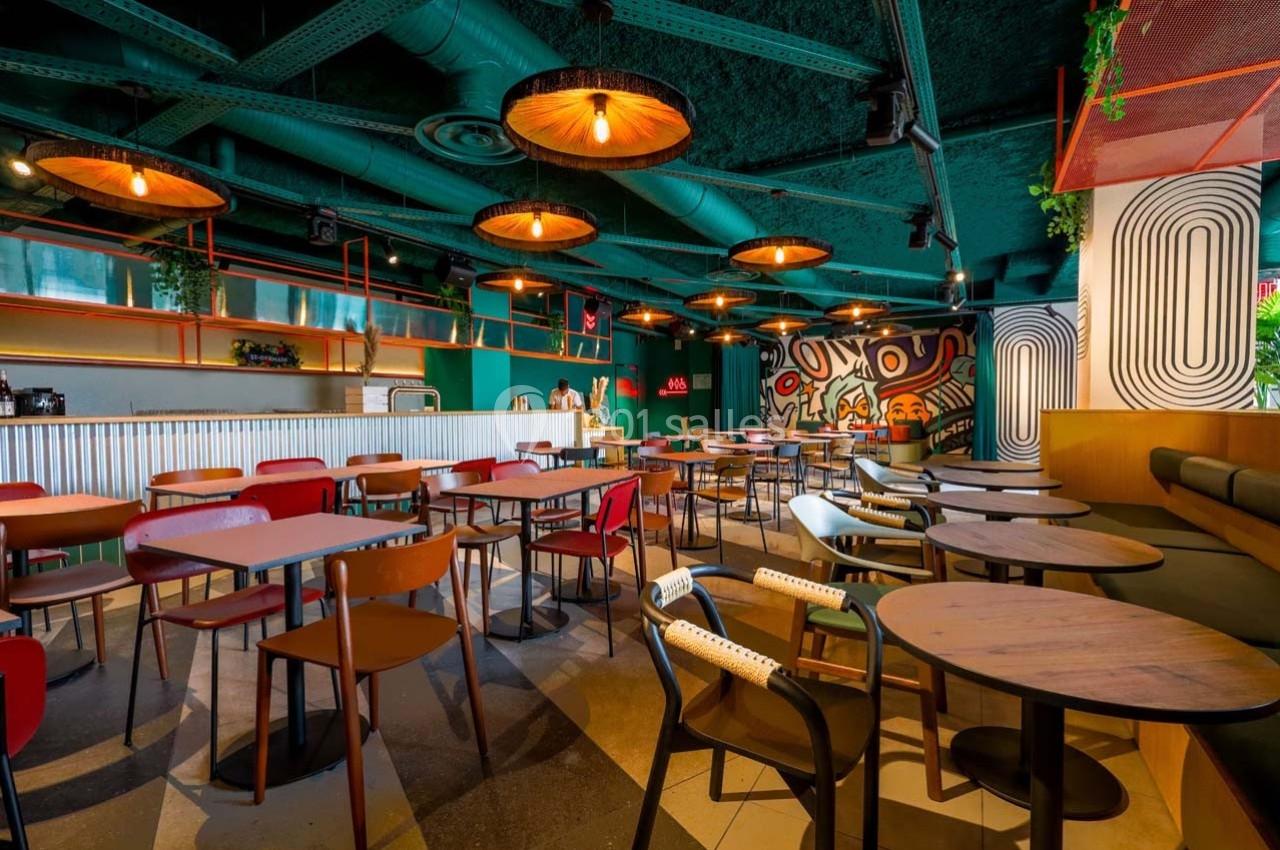 Salle de restaurant moderne avec tables en bois, chaises colorées, éclairage suspendu et mur décoré de motifs artistiques.