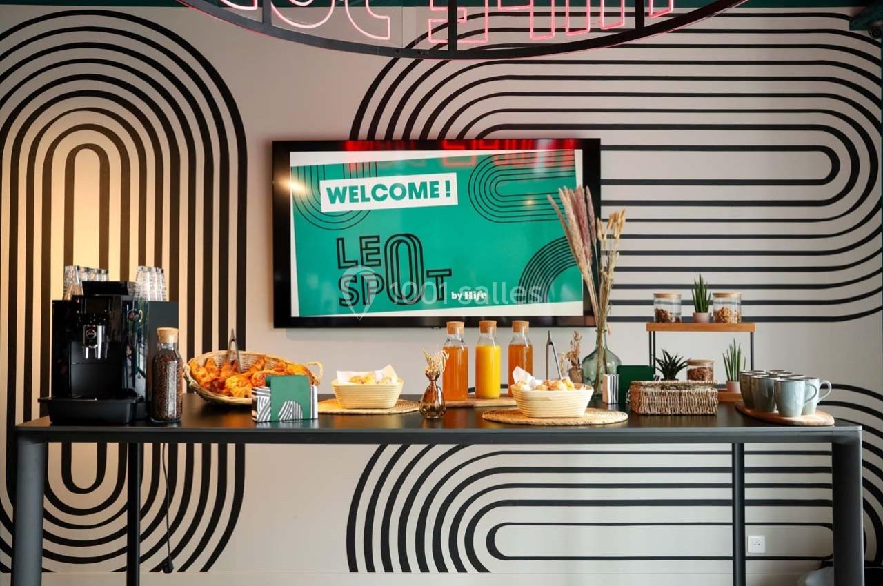 Table avec boissons, viennoiseries et décorations devant un mur graphique et un écran affichant ’Welcome! Le Spot’.