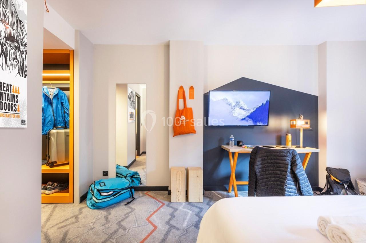 Chambre d'hôtel moderne avec lit, bureau, écran mural, valises et vêtements rangés dans un coin ouvert.