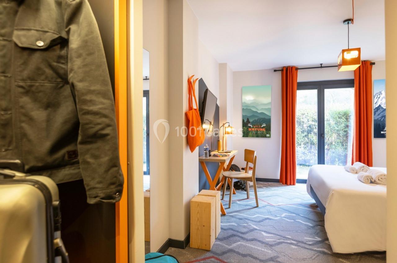 Chambre lumineuse avec lit double, bureau, rideaux orange et vue sur un jardin à travers une baie vitrée.