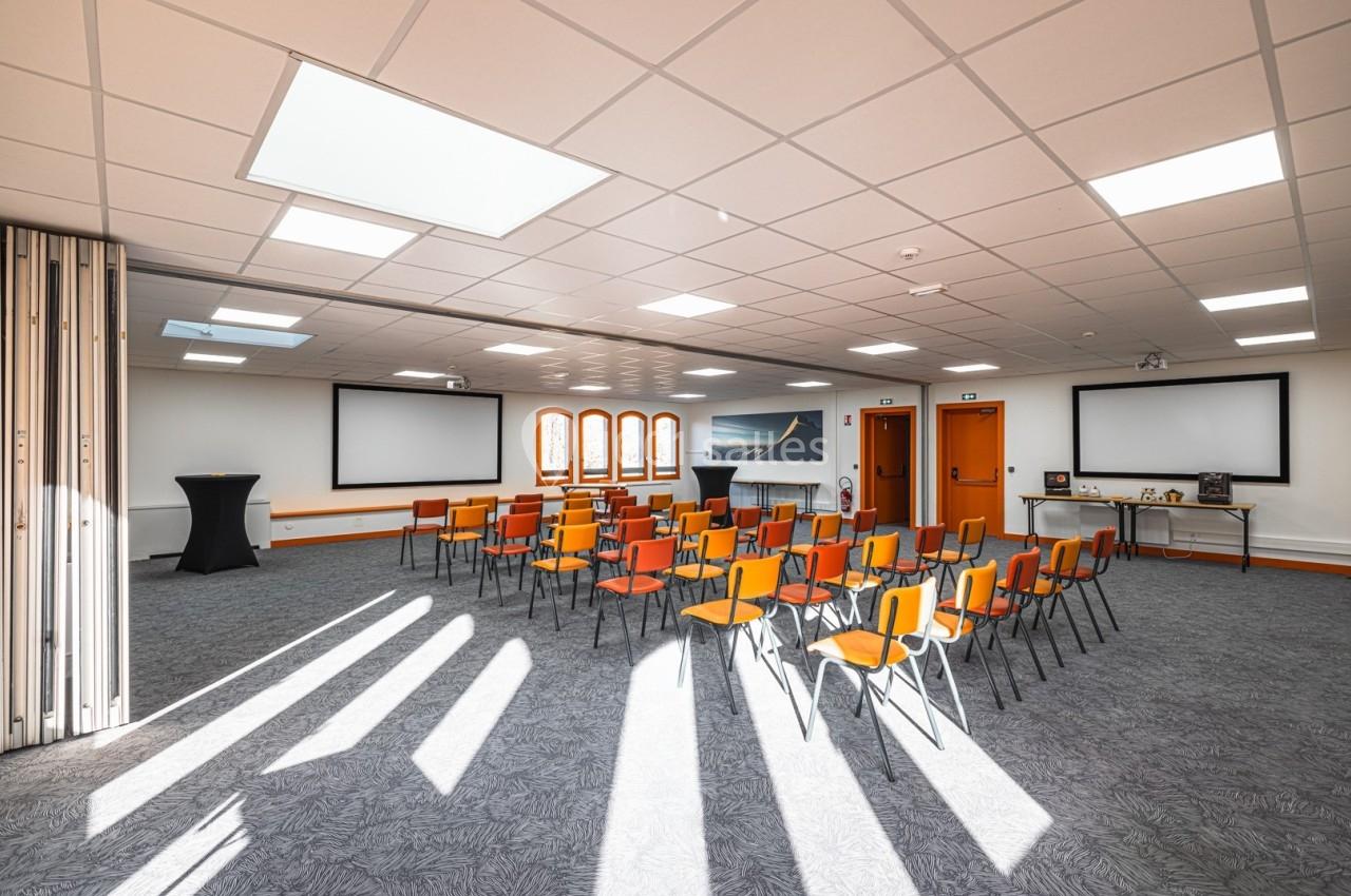 Salle de conférence lumineuse avec des chaises orange disposées en rangées, écrans et tables en arrière-plan.