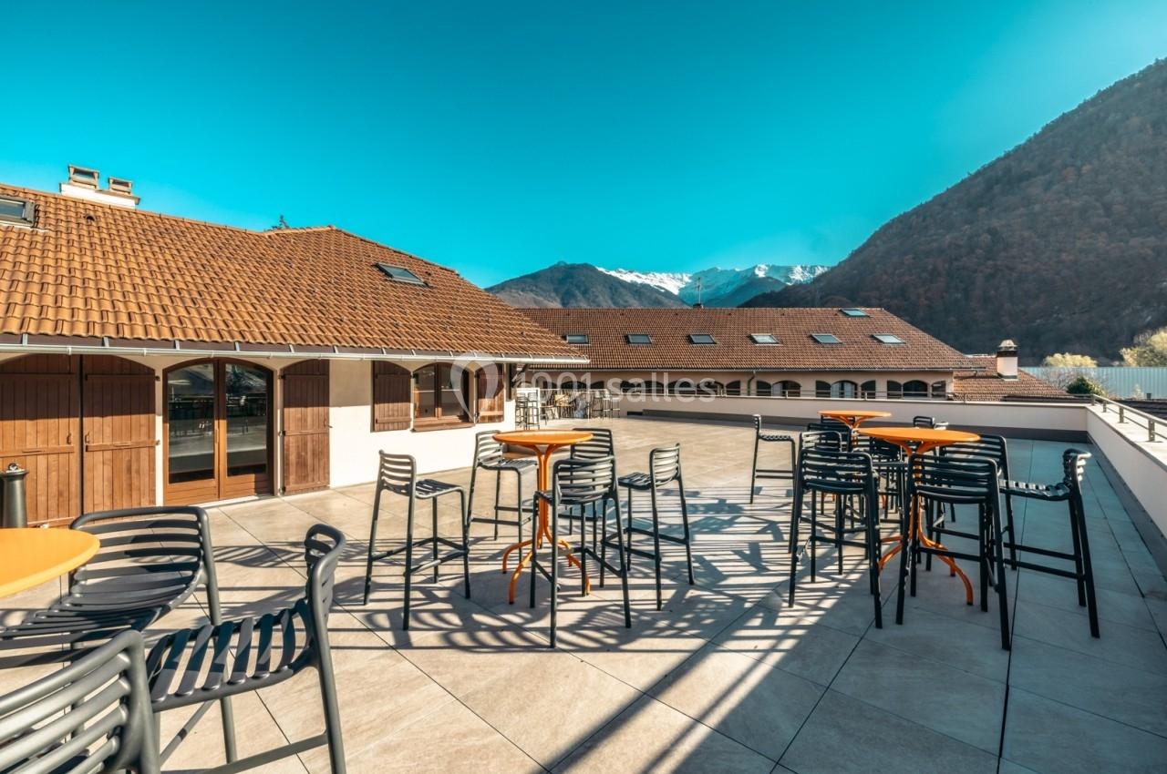 Terrasse ensoleillée avec tables et chaises, vue sur des montagnes enneigées et bâtiments aux toits en tuiles.