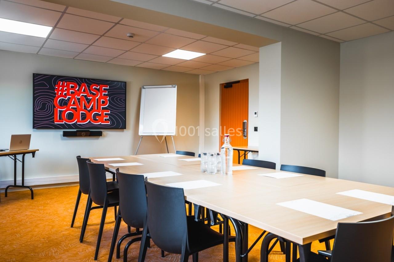 Salle de réunion lumineuse avec table rectangulaire, chaises noires, écran affichant ’Base Camp Lodge’ et tableau blanc.
