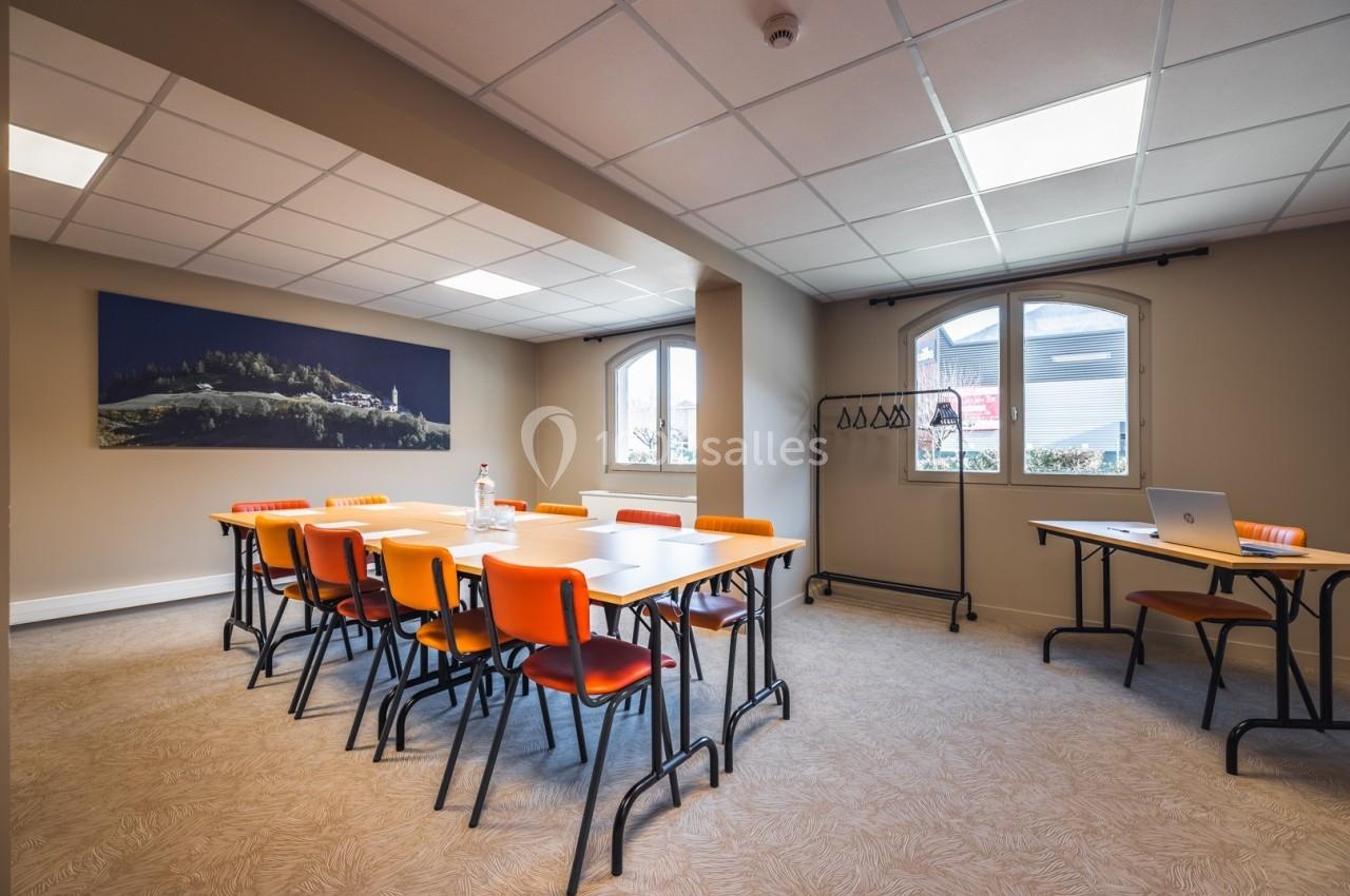 Salle de réunion lumineuse avec tables rectangulaires, chaises orange, porte-manteaux et vue sur l'extérieur par deux…