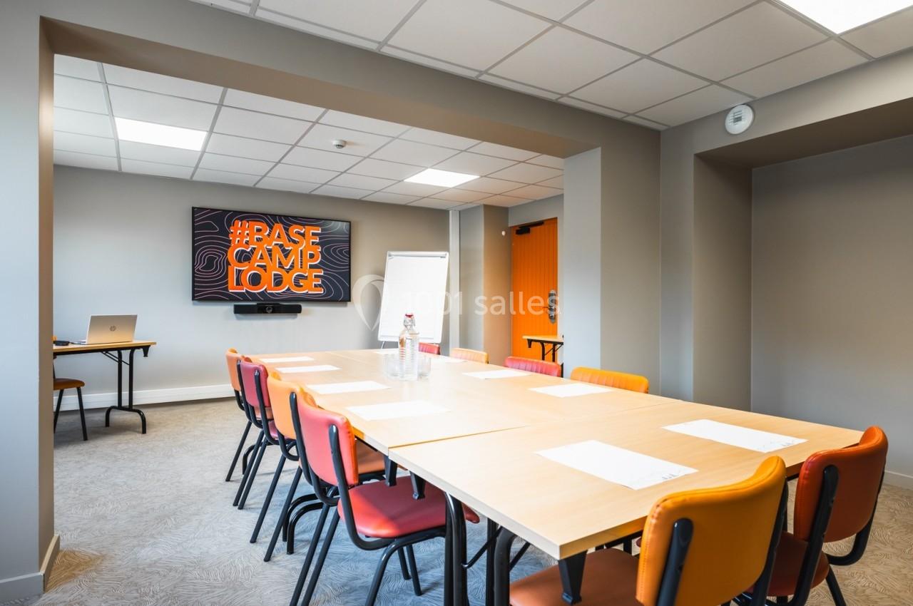 Salle de réunion lumineuse avec table rectangulaire, chaises colorées, écran mural, tableau blanc et bouteilles d'eau.