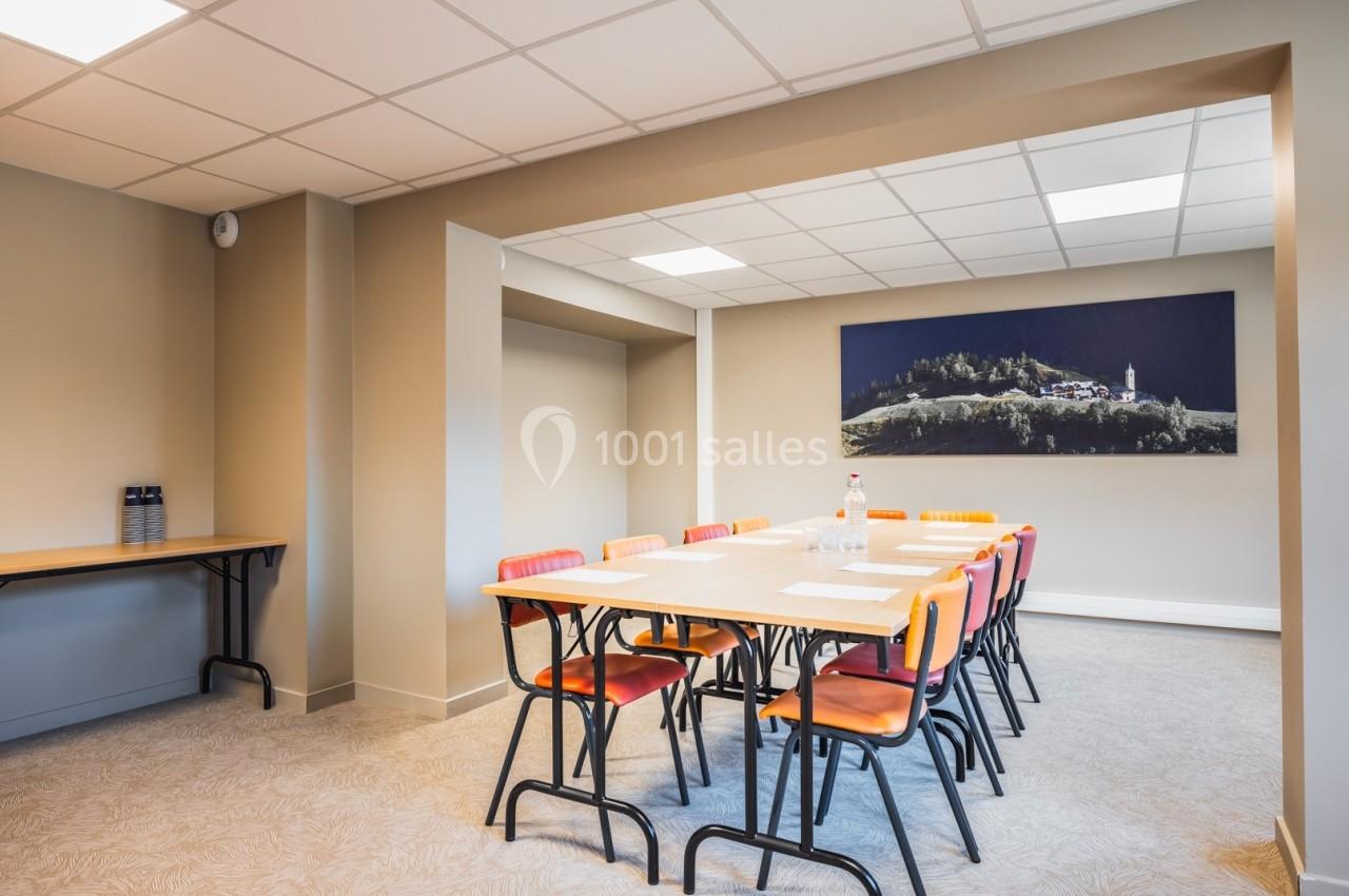 Salle de réunion lumineuse avec table rectangulaire, chaises colorées et tableau mural représentant un paysage.