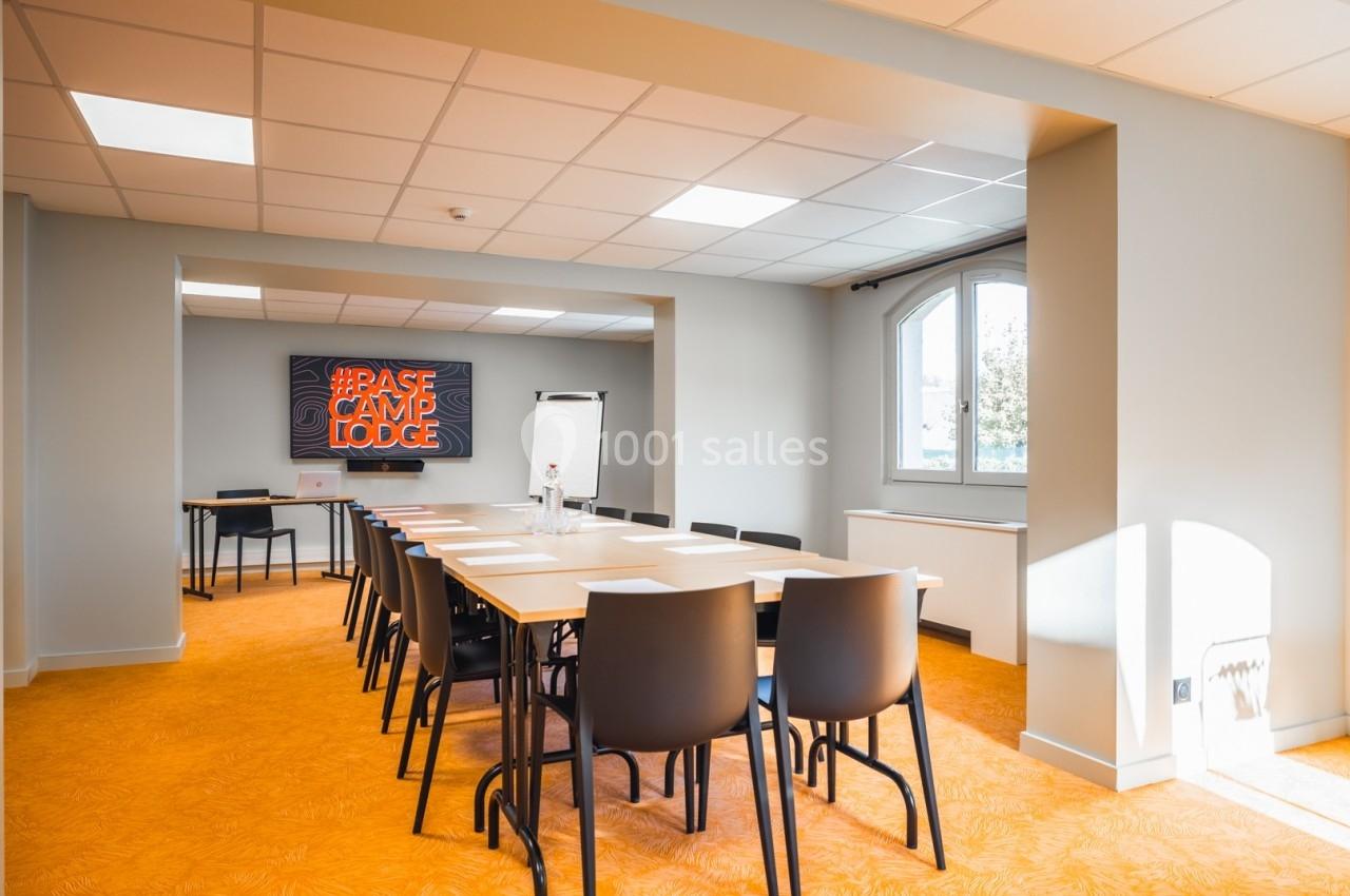 Salle de réunion lumineuse avec une grande table, des chaises noires, un tableau blanc et un écran affichant un néon orange.