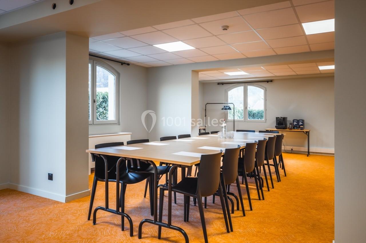 Salle de réunion lumineuse avec une grande table rectangulaire entourée de chaises noires, sol orange et fenêtres en arrière…
