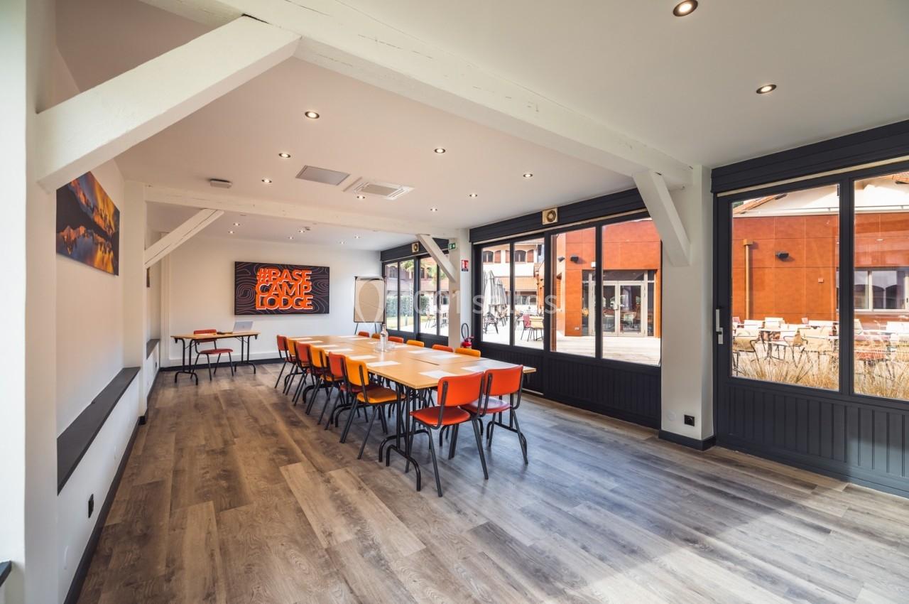 Salle lumineuse avec tables et chaises orange, grandes fenêtres donnant sur une terrasse et murs blancs avec décorations.