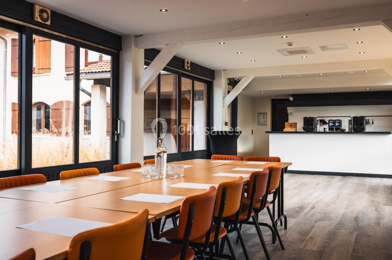 Salle de réunion lumineuse avec grande table, chaises orange, baies vitrées et coin café en arrière-plan.