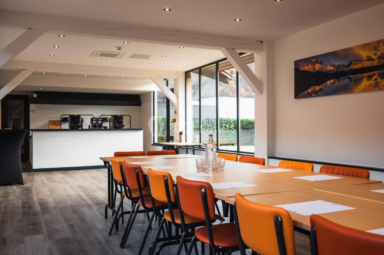 Salle de réunion lumineuse avec table en bois, chaises orange, baies vitrées et coin café en arrière-plan.