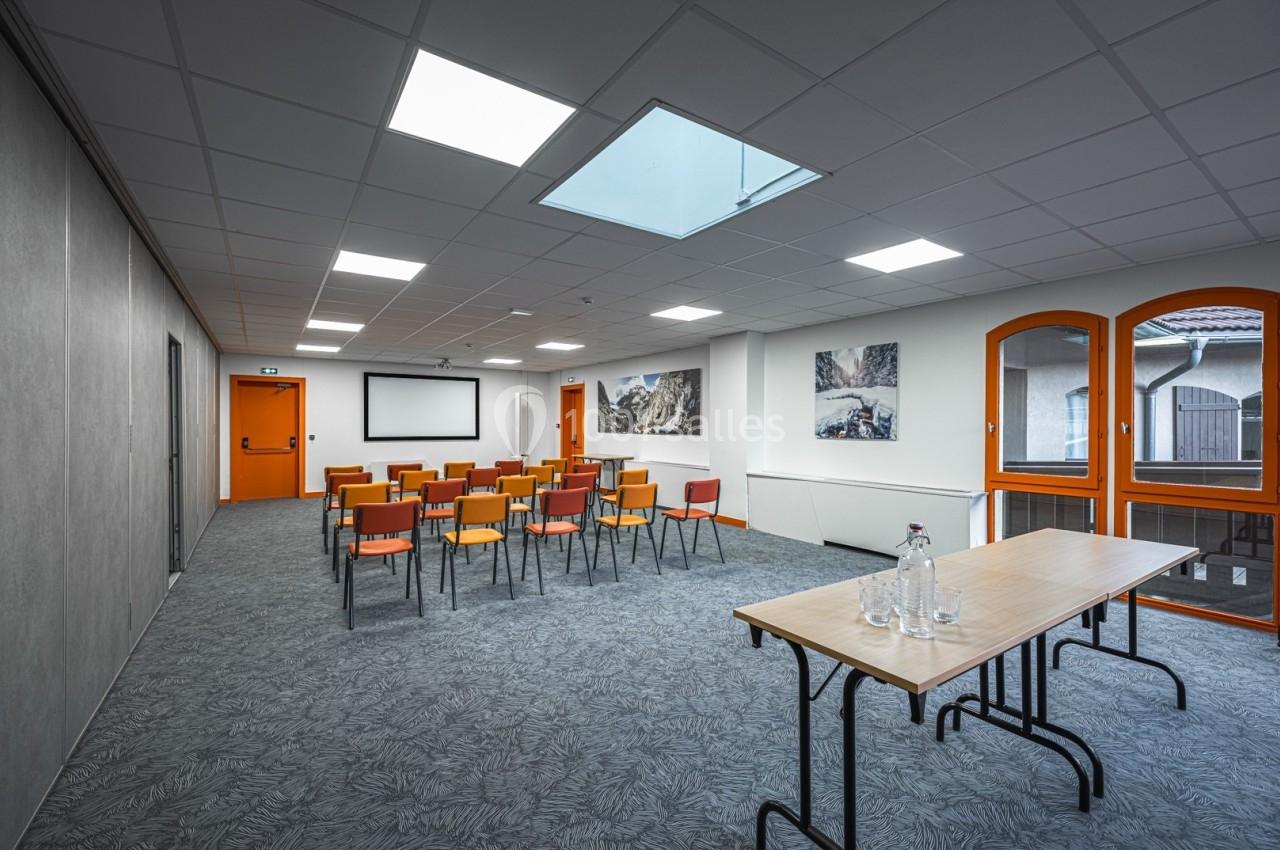 Salle de réunion lumineuse avec chaises alignées, écran de projection, table en bois et grandes fenêtres.