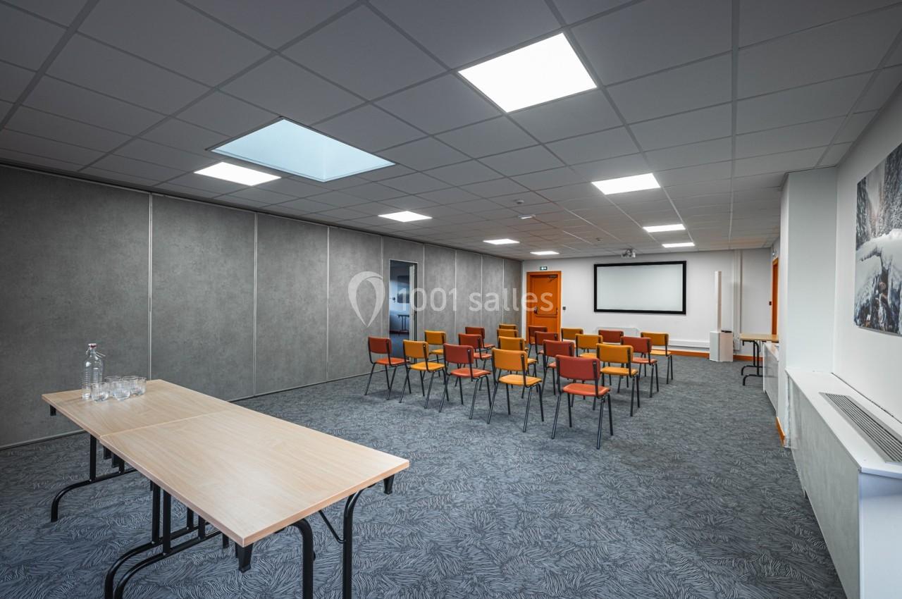 Salle de réunion avec chaises colorées alignées, écran de projection, tables en bois et éclairage au plafond.