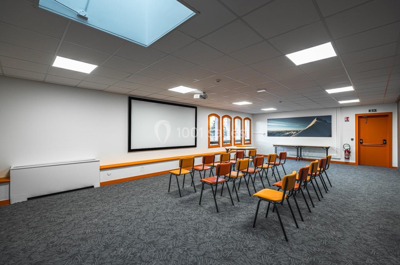 Salle de réunion lumineuse avec chaises alignées, écran de projection, fenêtres et éclairage au plafond.